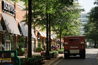 A shopping street with several storefronts, including a DSW Shoes store with black and white striped awnings. Trees line the walkway, providing shade. There is a red armored truck parked on the street and people can be seen walking or sitting on benches along the sidewalk. The surrounding area appears to be clean and well-maintained, with street lights and potted plants along the path.