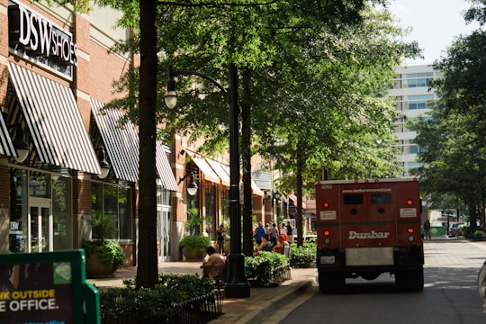 A shopping street with several storefronts, including a DSW Shoes store with black and white striped awnings. Trees line the walkway, providing shade. There is a red armored truck parked on the street and people can be seen walking or sitting on benches along the sidewalk. The surrounding area appears to be clean and well-maintained, with street lights and potted plants along the path.