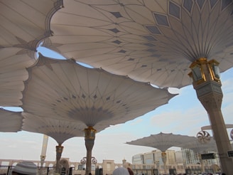 Large architectural umbrellas cover an outdoor area, supported by intricately designed columns with gold accents. The setting appears to be a public square or plaza, with buildings visible in the distance under a clear blue sky.