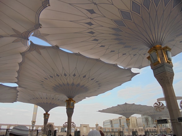 Large architectural umbrellas cover an outdoor area, supported by intricately designed columns with gold accents. The setting appears to be a public square or plaza, with buildings visible in the distance under a clear blue sky.