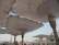 Large architectural umbrellas cover an outdoor area, supported by intricately designed columns with gold accents. The setting appears to be a public square or plaza, with buildings visible in the distance under a clear blue sky.