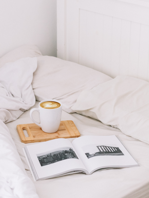 A cozy bed setup featuring a cup of coffee on a wooden tray and an open book displaying black and white photographs. The soft bedding and natural light create a relaxed atmosphere.