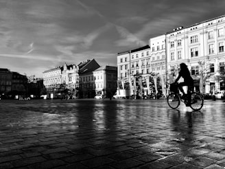 A black and white photograph featuring a person riding a bicycle across a cobblestone square. The background consists of historic buildings with intricate architectural details. The lighting creates stark contrasts and long shadows, adding drama to the scene.