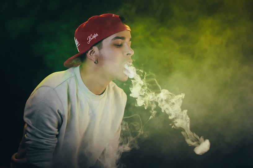 A person wearing a red cap and a light-colored sweatshirt is blowing smoke rings. The background is filled with dense smoke with shades of green.