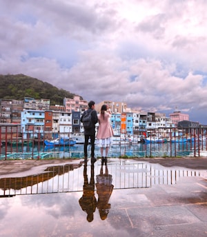 A couple stands on a waterfront with their reflections visible in a puddle. They overlook a colorful row of buildings and boats under a cloudy sky.