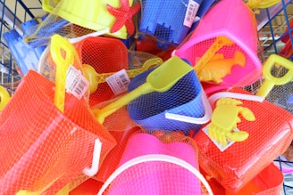 A collection of brightly colored plastic beach toys, including shovels, buckets, and crab and starfish molds, all wrapped in netting. The toys are in shades of yellow, red, blue, and pink, and appear to be packed in a basket or display.