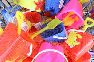 A collection of brightly colored plastic beach toys, including shovels, buckets, and crab and starfish molds, all wrapped in netting. The toys are in shades of yellow, red, blue, and pink, and appear to be packed in a basket or display.