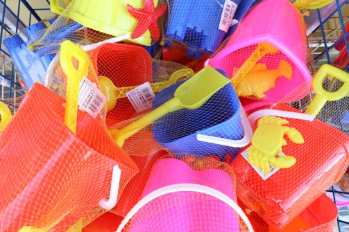 A collection of brightly colored plastic beach toys, including shovels, buckets, and crab and starfish molds, all wrapped in netting. The toys are in shades of yellow, red, blue, and pink, and appear to be packed in a basket or display.
