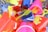 A collection of brightly colored plastic beach toys, including shovels, buckets, and crab and starfish molds, all wrapped in netting. The toys are in shades of yellow, red, blue, and pink, and appear to be packed in a basket or display.