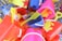 A collection of brightly colored plastic beach toys, including shovels, buckets, and crab and starfish molds, all wrapped in netting. The toys are in shades of yellow, red, blue, and pink, and appear to be packed in a basket or display.