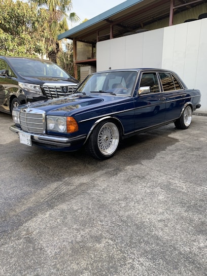 A classic blue sedan with a vintage design is parked on a concrete surface. It has wire-spoke wheels and prominent chrome detailing on the grille and bumper. Next to it is a modern black minivan with a sleek design. The background features green foliage and a building with a metal roof.
