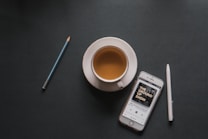 A cup of coffee sits on a dark surface accompanied by a smartphone displaying a podcast titled 'The Ground Up Show.' A pencil and a pen are positioned nearby, creating a balanced and minimalistic layout.