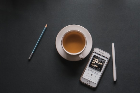 A cup of coffee sits on a dark surface accompanied by a smartphone displaying a podcast titled 'The Ground Up Show.' A pencil and a pen are positioned nearby, creating a balanced and minimalistic layout.