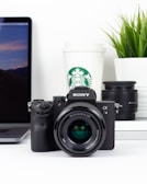 A Sony camera is placed on a desk in front of a laptop with a sunset wallpaper. To the right, there's a Starbucks cup and a plant with green leaves. Another camera lens is visible next to some books.