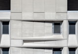 A concrete facade featuring a geometric arrangement of rectangular panels. Three dark, narrow windows are spaced evenly across the structure. A mesh-covered window is positioned slightly off-center in the upper part. The design emphasizes a modernist aesthetic with clean lines and shadow play across the surface.