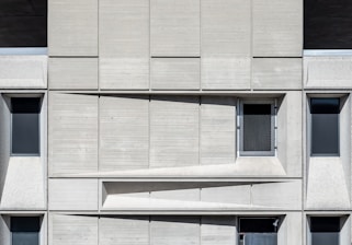 A concrete facade featuring a geometric arrangement of rectangular panels. Three dark, narrow windows are spaced evenly across the structure. A mesh-covered window is positioned slightly off-center in the upper part. The design emphasizes a modernist aesthetic with clean lines and shadow play across the surface.