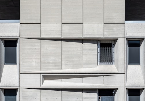 A concrete facade featuring a geometric arrangement of rectangular panels. Three dark, narrow windows are spaced evenly across the structure. A mesh-covered window is positioned slightly off-center in the upper part. The design emphasizes a modernist aesthetic with clean lines and shadow play across the surface.