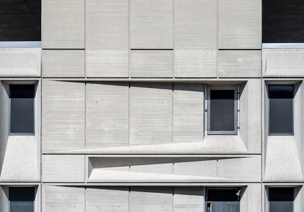 A concrete facade featuring a geometric arrangement of rectangular panels. Three dark, narrow windows are spaced evenly across the structure. A mesh-covered window is positioned slightly off-center in the upper part. The design emphasizes a modernist aesthetic with clean lines and shadow play across the surface.