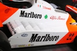 The side view of a Formula 1 racing car prominently featuring branding such as Marlboro, Alfa Romeo, and Michelin. The car has striking orange and white colors, with a visible Italian flag and a four-leaf clover symbol near the driver's name, B. Giacomelli.