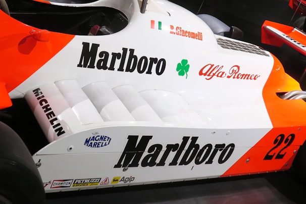 The side view of a Formula 1 racing car prominently featuring branding such as Marlboro, Alfa Romeo, and Michelin. The car has striking orange and white colors, with a visible Italian flag and a four-leaf clover symbol near the driver's name, B. Giacomelli.