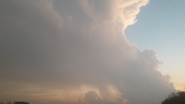A large, dramatic cloud formation dominates the sky, with the sunlight creating subtle hues in the background, ranging from light blue to soft orange. The contrast between the dark, dense clouds and the lighter sky gives a sense of impending weather change or storm.