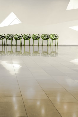A row of seven green, intricately designed chairs is aligned against a plain beige wall with angular white windows. The glossy tiled floor reflects the chairs and windows, creating a calm and symmetrical appearance.