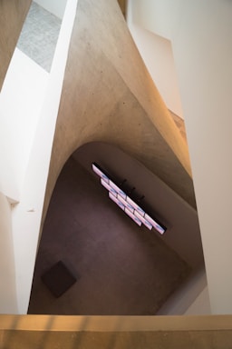 A modern architectural space with angular, concrete walls and a multi-screen video installation on the opposite side. The minimalist interior creates a sense of depth and perspective.