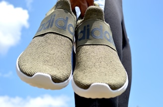 A pair of olive-green slip-on sneakers with white soles held against a bright blue sky background. The sneakers feature a prominent brand logo on the strap across the top.