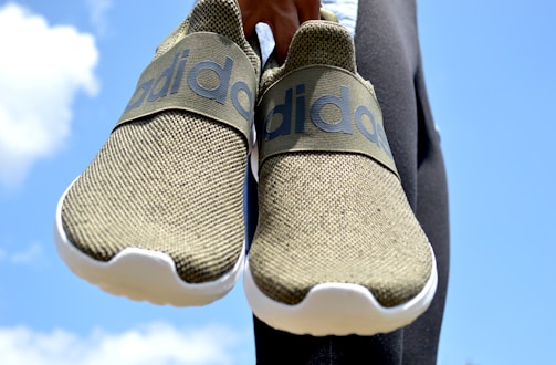 A pair of olive-green slip-on sneakers with white soles held against a bright blue sky background. The sneakers feature a prominent brand logo on the strap across the top.