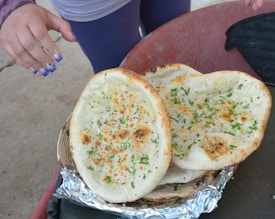 A close-up view of flatbreads topped with herbs and black seeds, resting on aluminum foil. A person's hand with decorated fingernails is reaching towards the bread. The flatbreads are slightly browned and garnished, resting on a wicker basket.