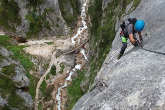 A person wearing climbing gear and a helmet is scaling a steep rock face using metal rungs and a safety cable. Below, a winding, narrow stream runs through a rocky gorge, with lush greenery lining the path. A wooden bridge crosses the stream, and the overall scene is set in a mountainous landscape.