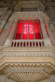 An ornate building facade featuring a large window with a red neon letter 'R' inside. The architectural elements include sculpted columns and intricate stone details, suggesting a classical or historic style.