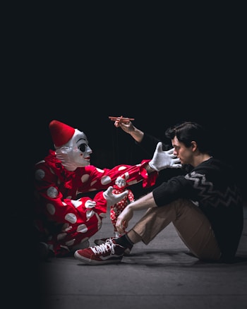 A person dressed as a clown in a red and white polka-dot costume and a red hat is crouched on the ground, interacting with a man sitting cross-legged. The clown is playfully controlling a small puppet dressed similarly with strings. The scene is set against a dark background, providing a mysterious and theatrical atmosphere.
