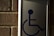 A metal sign with a blue symbol of a person in a wheelchair, mounted on a brick wall.
