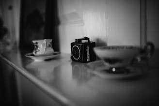 A black and white photograph featuring a vintage camera placed on a reflective surface beside ornate teacups on saucers, creating a nostalgic and classic atmosphere.