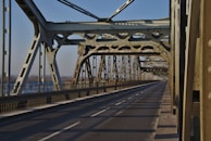 A metal bridge structure with intricate steel beams and a long, empty road extending into the distance. The bridge design features cross-bracing and riveted joints, showcasing industrial architecture. Sunlight casts shadows across the road surface, creating a pattern of light and shadow.