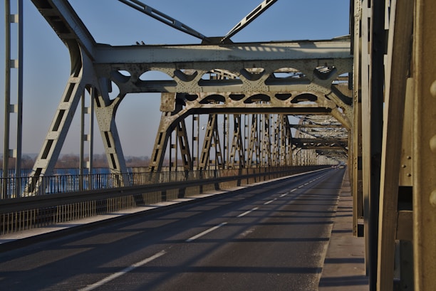 A metal bridge structure with intricate steel beams and a long, empty road extending into the distance. The bridge design features cross-bracing and riveted joints, showcasing industrial architecture. Sunlight casts shadows across the road surface, creating a pattern of light and shadow.