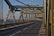 A metal bridge structure with intricate steel beams and a long, empty road extending into the distance. The bridge design features cross-bracing and riveted joints, showcasing industrial architecture. Sunlight casts shadows across the road surface, creating a pattern of light and shadow.