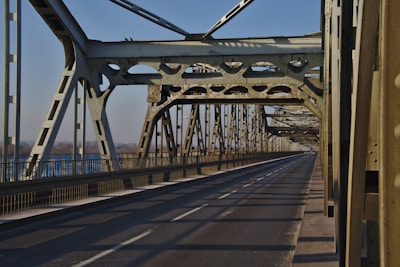 A metal bridge structure with intricate steel beams and a long, empty road extending into the distance. The bridge design features cross-bracing and riveted joints, showcasing industrial architecture. Sunlight casts shadows across the road surface, creating a pattern of light and shadow.