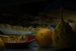 A dimly lit scene with a quiche slice on a red plate, accompanied by a dark, bumpy yellow gourd, an orange, and another quiche in the background. The arrangement suggests a fresh and rustic culinary theme.