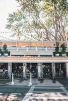 An outdoor view of a restaurant called Sunset Beach, featuring orange and white striped awnings. The setting includes trees with lanterns hanging from branches above. Seating is visible inside with open doors, and several plants are arranged around the restaurant.