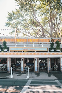 An outdoor view of a restaurant called Sunset Beach, featuring orange and white striped awnings. The setting includes trees with lanterns hanging from branches above. Seating is visible inside with open doors, and several plants are arranged around the restaurant.