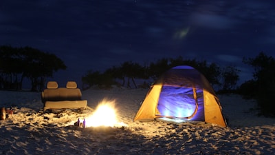 A nighttime camping scene on a sandy beach with a lit campfire and a brightly illuminated tent. An abandoned car seat is placed near the fire. The surrounding area is dark, with silhouettes of trees in the background, highlighting a peaceful and secluded setting.