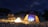 Campfire scene with tents set up near a calm beach under starry sky.