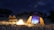 Campfire scene with tents set up near a calm beach under starry sky.