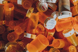 Numerous orange prescription pill bottles are piled together, with some displaying readable labels and white caps. The scene is cluttered and busy, evoking a sense of disarray.