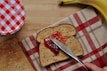 A slice of whole-grain bread is being spread with red jam using a knife. Beside it, there is another slice of toasted bread on a red and white checkered tea towel. A banana is placed in the upper right corner, and a red and white checkered jar lid is partially visible on the upper left side.