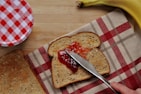 A slice of whole-grain bread is being spread with red jam using a knife. Beside it, there is another slice of toasted bread on a red and white checkered tea towel. A banana is placed in the upper right corner, and a red and white checkered jar lid is partially visible on the upper left side.