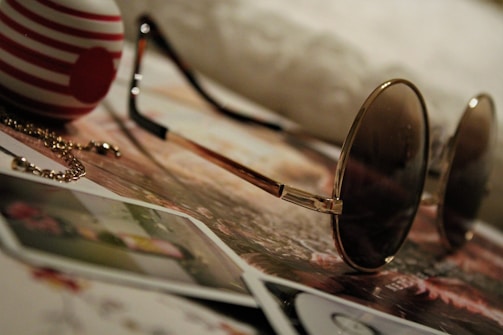 A pair of round sunglasses rests atop a collection of photographs and papers. Nearby, a striped object, possibly an egg-shaped ornament, adds a touch of color with its red and white pattern. A delicate chain lies on the surface alongside these items, creating a scene of casual elegance.