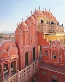 An iconic palace featuring intricate pink sandstone architecture with multiple arched windows and domes. Several people are observing the view from a balcony. The structure showcases traditional craftsmanship with detailed patterns and embellishments.